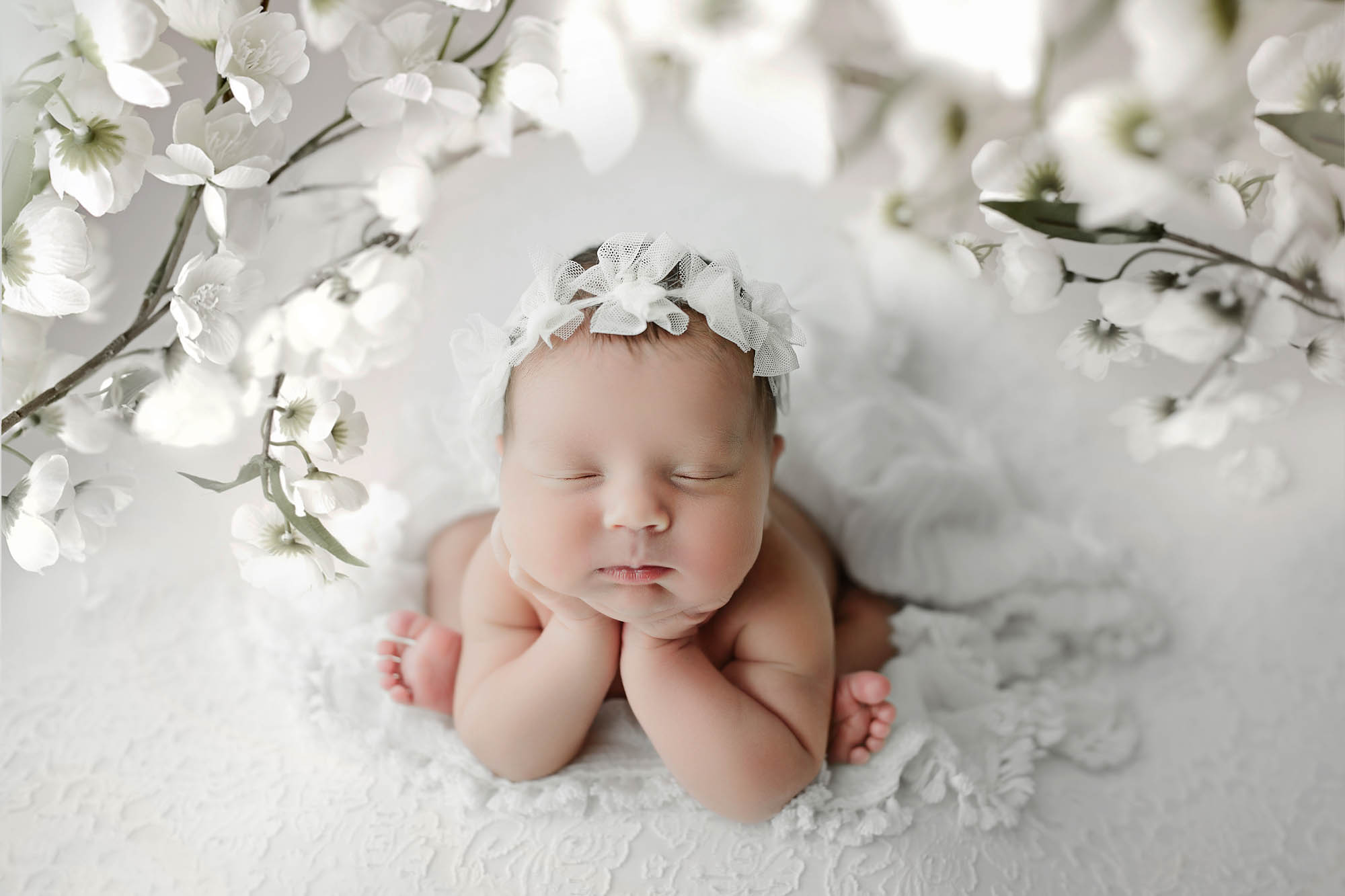 Canton Newborn Photographer image of newborn baby girl wearing a white floral headband with white flowers behind her laying on a white blanket with her hands holding her head